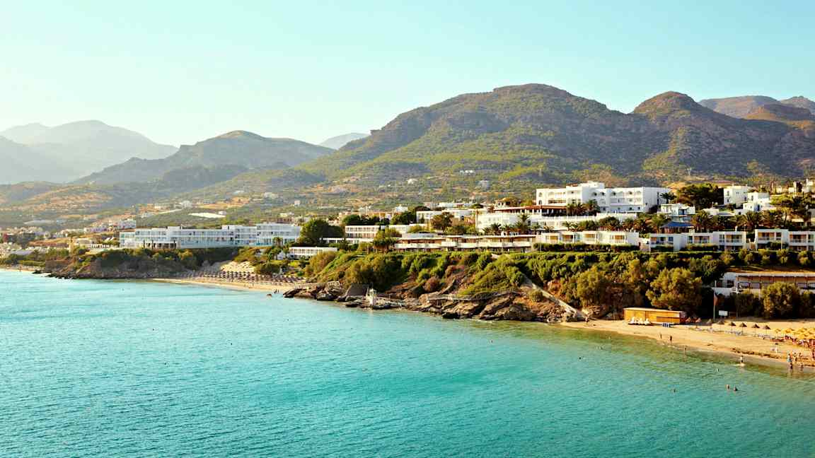 Flere bygninger nær en kyst med blått hav og fjell i bakgrunnen. (AI-generert tekst)