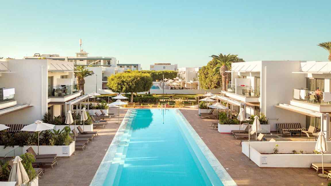 Et utendørsbasseng omgitt av moderne, hvite bygninger med balkonger og solsenger under parasoller. (AI-generert tekst)