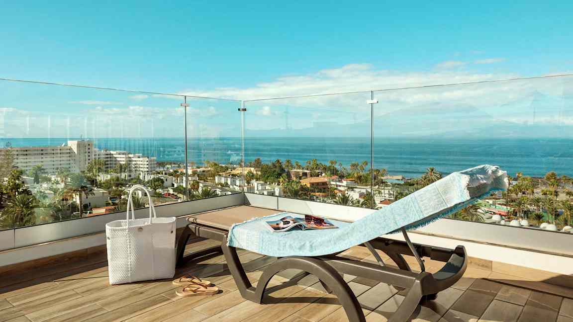 Utsikt fra en terrasse med en solseng dekket med et håndkle, en hvit veske og sandaler, over man ser havet og bygninger. (AI-generert tekst)