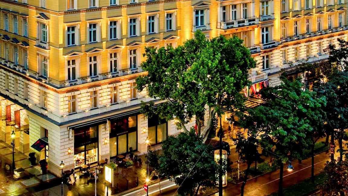 En opplyst hotellbygning med fasade i klassisk arkitektur og omkringliggende grøntområde og bymiljø. (AI-generert tekst)