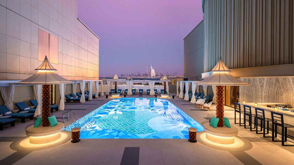 En takterrasse med et dekorativt basseng omgitt av solsenger og små parasoller. I bakgrunnen sees Burj Al Arab og Dubais skyline under en lilla kveldshimmel. (AI-generert tekst)