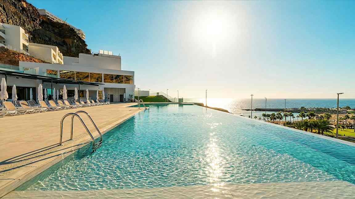 Et infinity-basseng med speilklart vann, solsenger og parasoller på siden, i et hotellområde med utsikt mot havet og stranden. (AI-generert tekst)