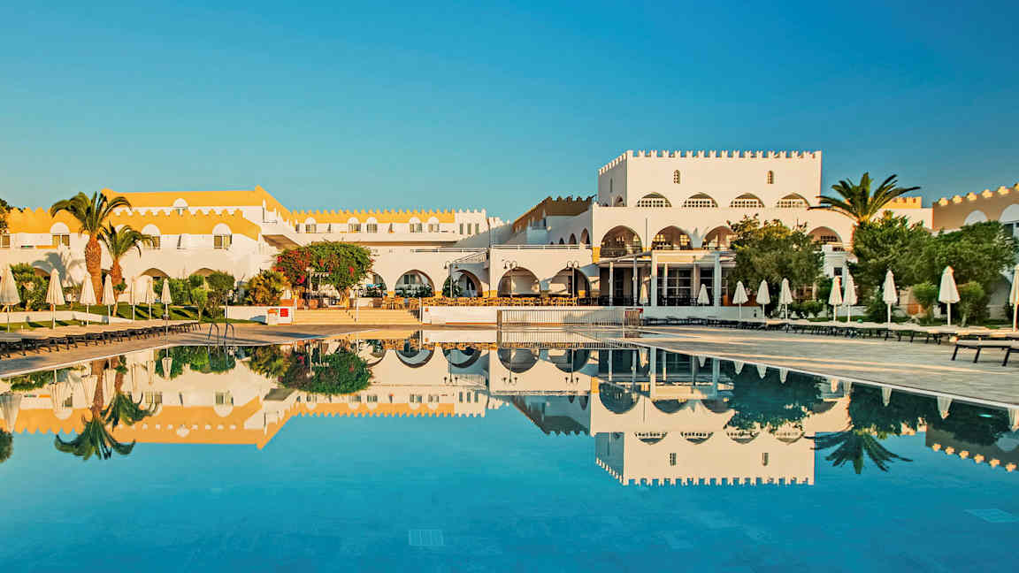 En hvit bygning med gult tak ved et basseng med solsenger og parasoller. (AI-generert tekst)