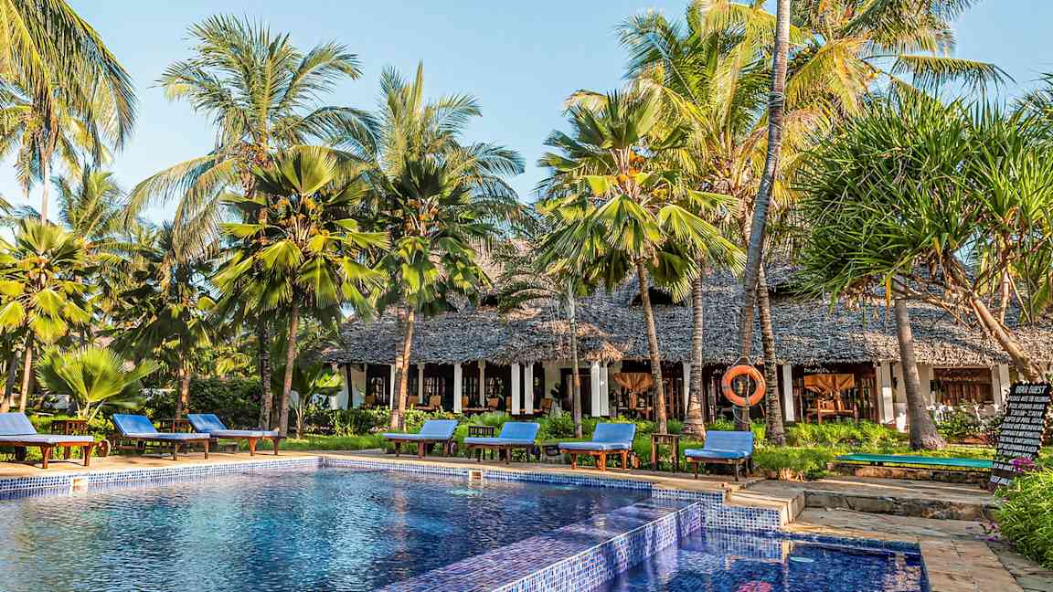 Et basseng omgitt av solsenger og palmer ved en bygning med stråtak. (AI-generert tekst)