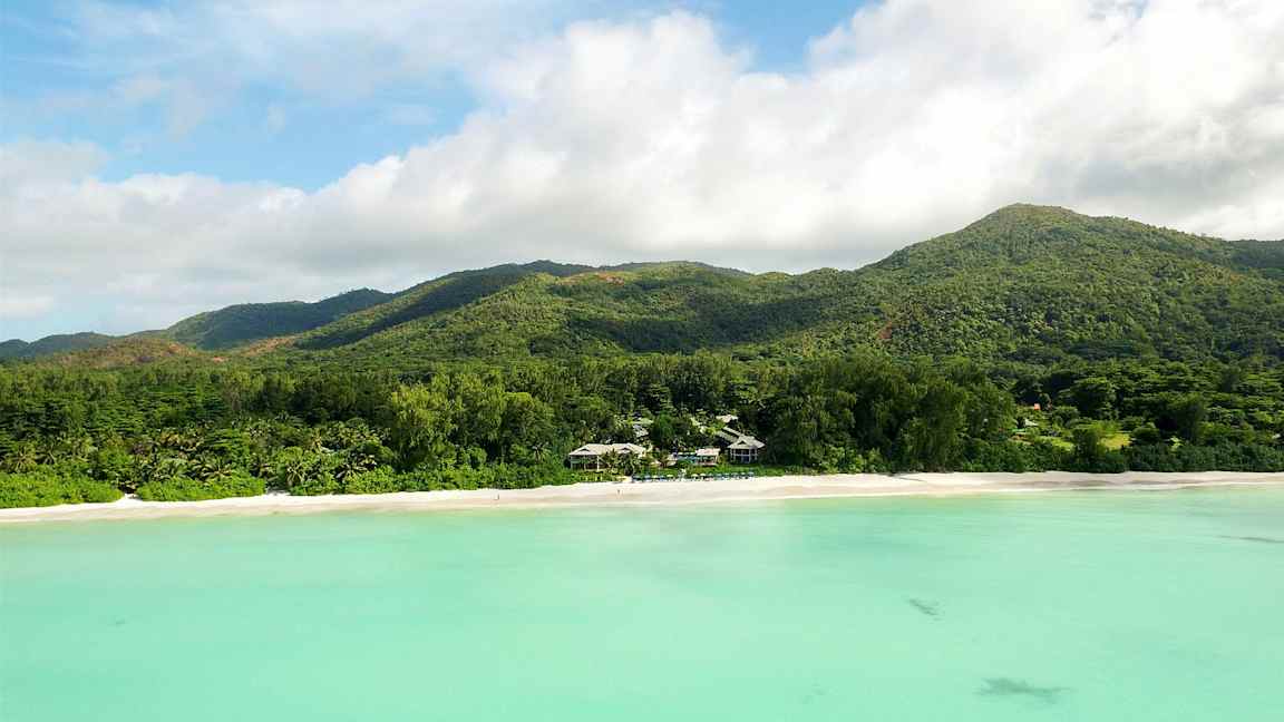 En strand med hvit sand foran en skogkledd fjellkjede og turkis vann. (AI-generert tekst)