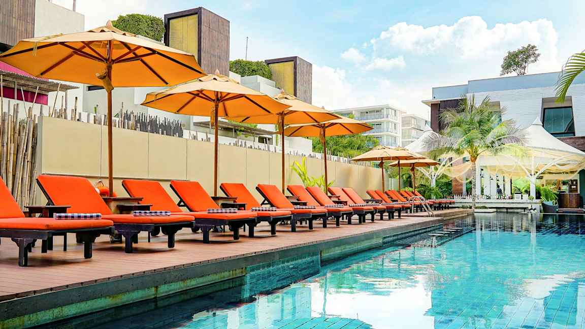 Et utendørsbasseng med oransje solsenger og parasoller ved kanten, omgitt av moderne arkitektur. (AI-generert tekst)