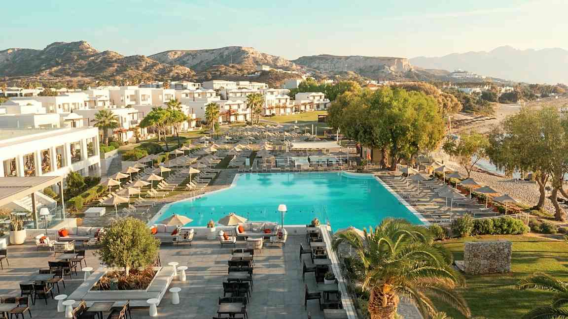 Flyfoto av en ferieanlegg med basseng, solsenger, bygninger og omkringliggende landskap. (AI-generert tekst)