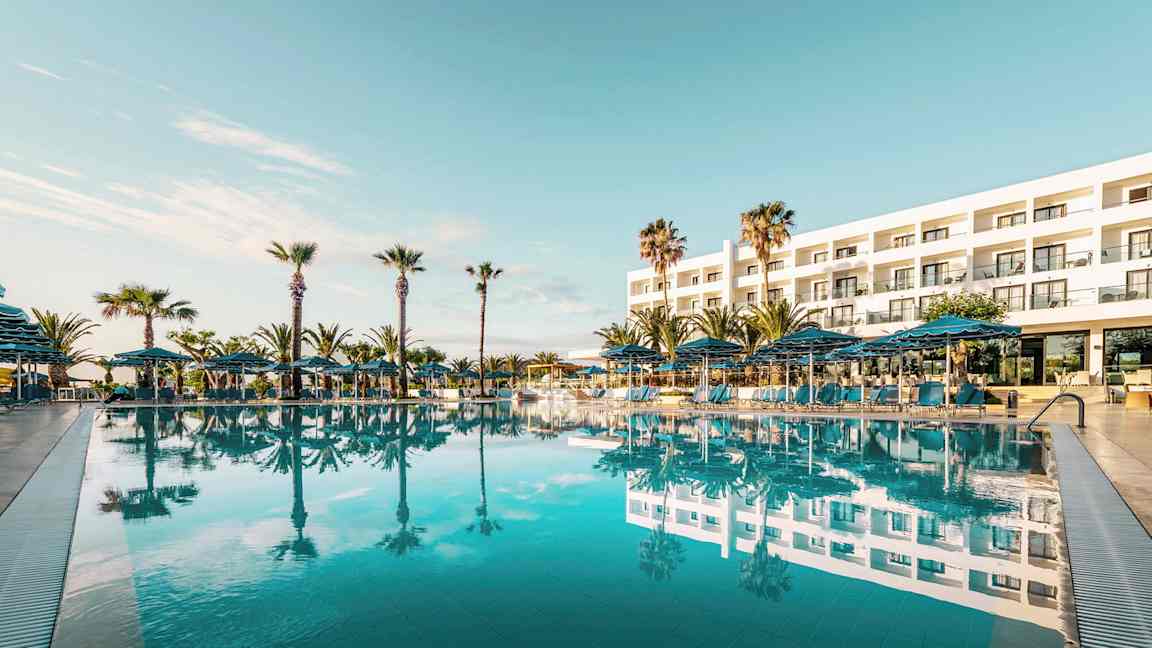 Eksteriør av et hotell med et stort basseng foran og omkransende palmer. (AI-generert tekst)