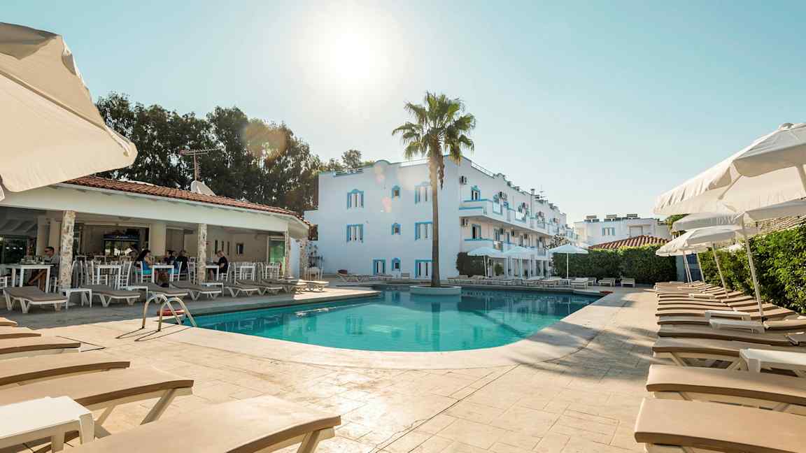 Bilde av et basseng med solsenger og parasoller, bak ses en hvit bygning og et åpent reservasjonsområde. (AI-generert tekst)