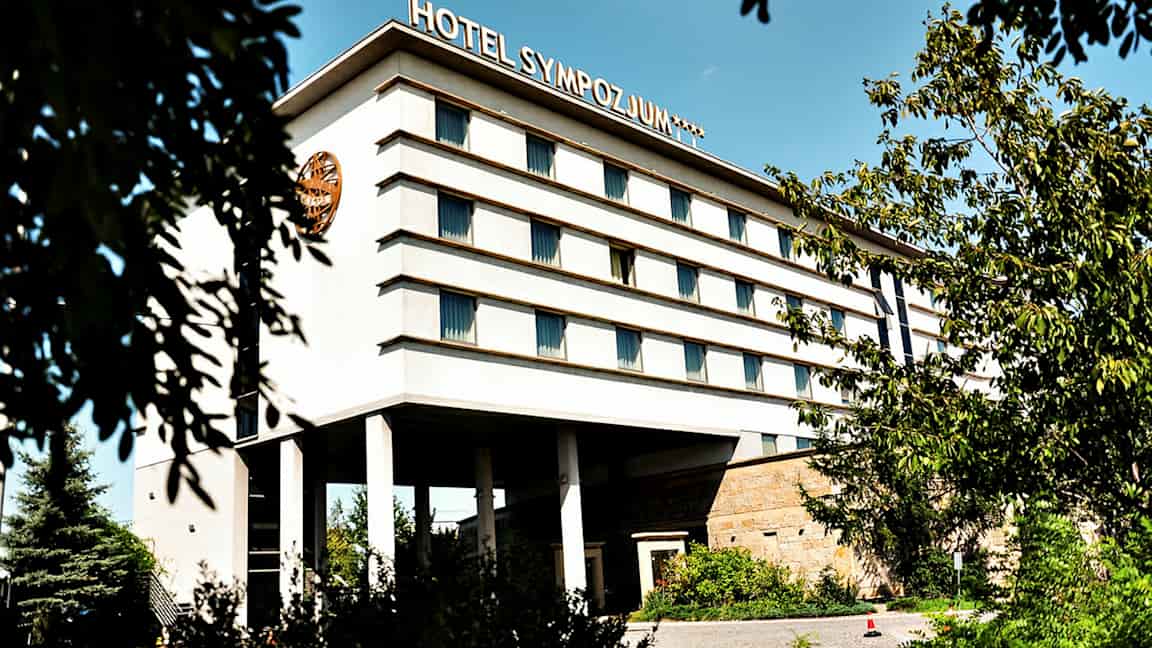 Bygning med flere etasjer, merket med teksten 'Hotel Sympozjum', omgitt av trær og vegetasjon. (AI-generert tekst)