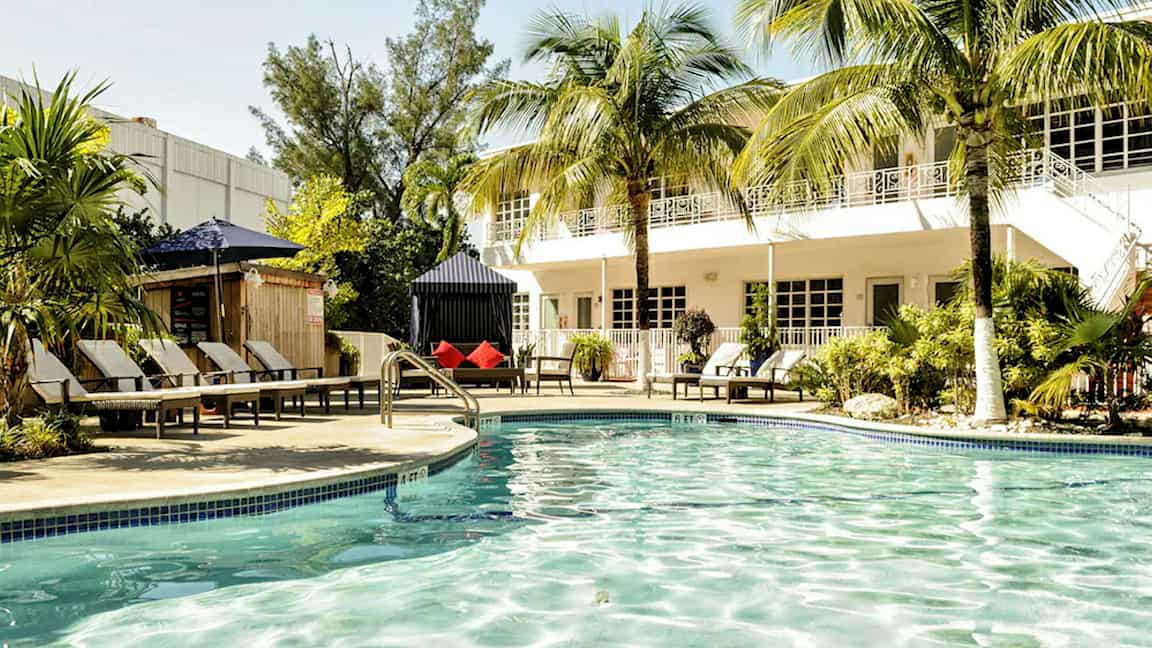 Et utendørs basseng omgitt av stoler, palmer og en bygning i bakgrunnen. (AI-generert tekst)