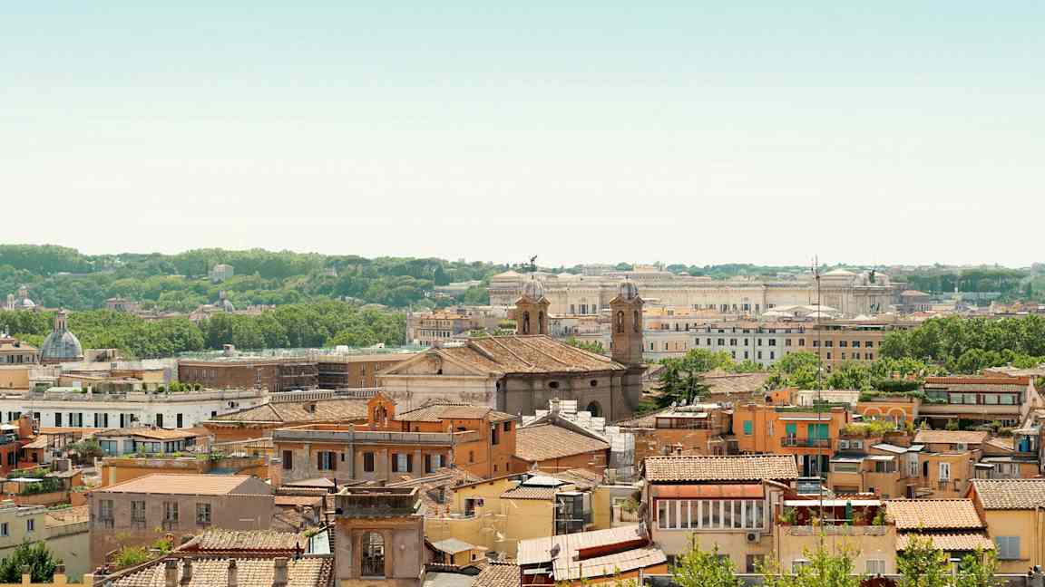 Utsikt over en by med mange bygninger, to kirketårn og grønn vegetasjon i bakgrunnen. (AI-generert tekst)