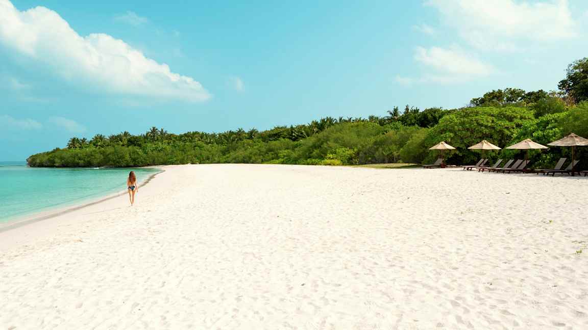 En kvinne går på en sandstrand med turkis vann og grønn vegetasjon i bakgrunnen. (AI-generert tekst)