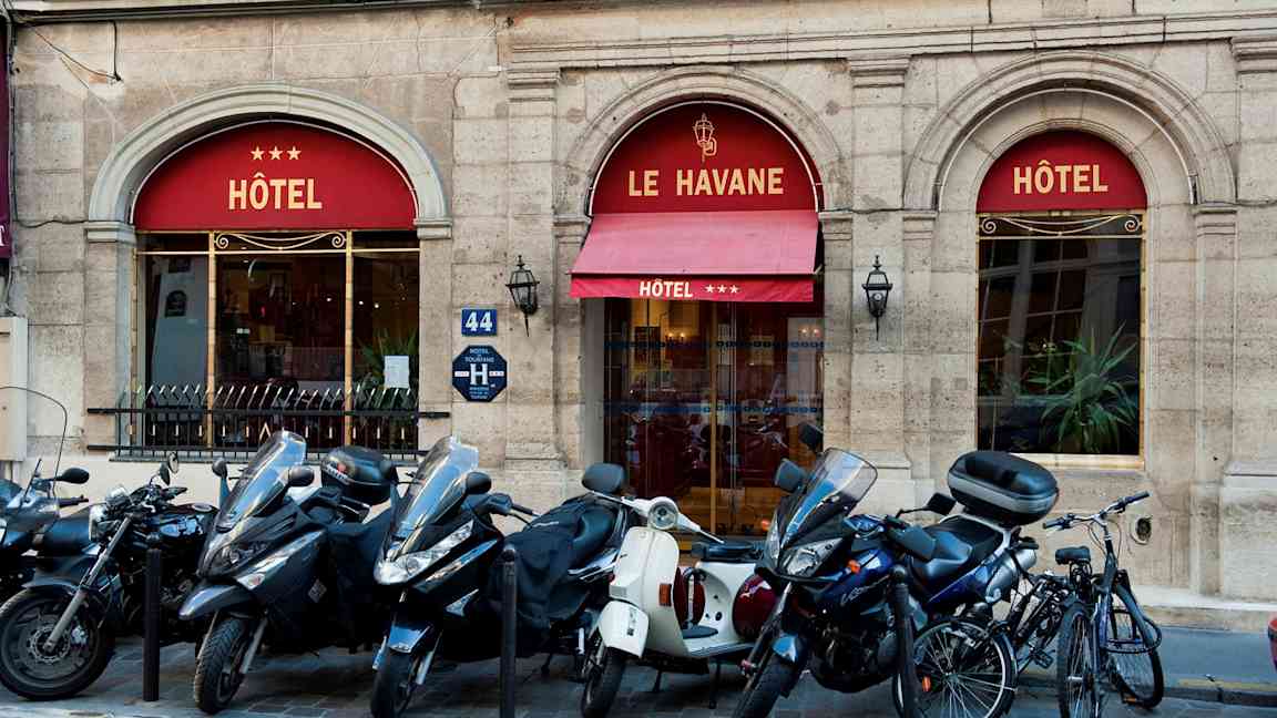 Bilde som viser inngangen til et hotell med navnet Le Havane, tre stjerner vises på markisen. Foran bygningen står mopeder og sykler parkert. (AI-generert tekst)