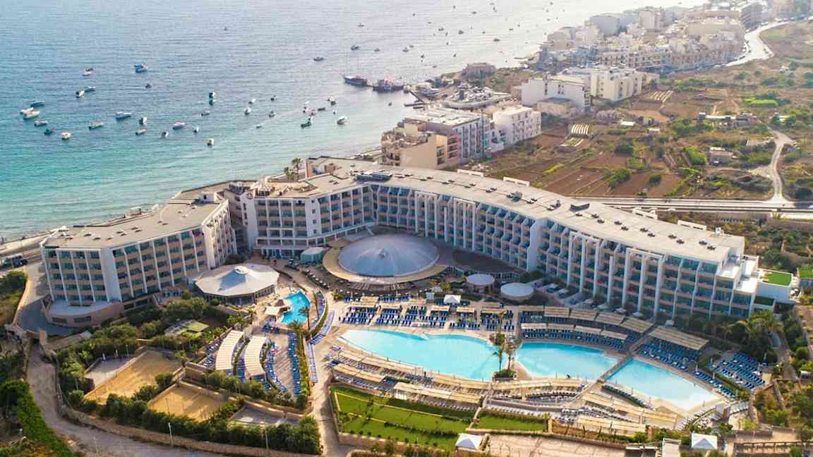 Luftfoto av et hotellkompleks med bassenger, grøntområder og havutsikt. (AI-generert tekst)