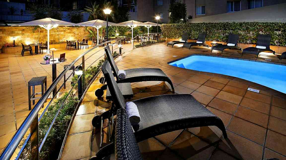 Hotellens bassengområde med solsenger og parasoller ved et opplyst flisebelagt uteområde. (AI-generert tekst)