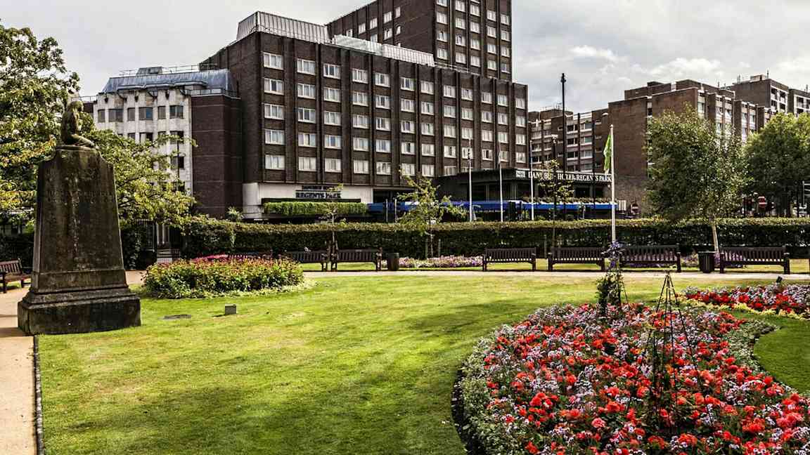 En park med blomster og gressplen foran en bygning. (AI-generert tekst)