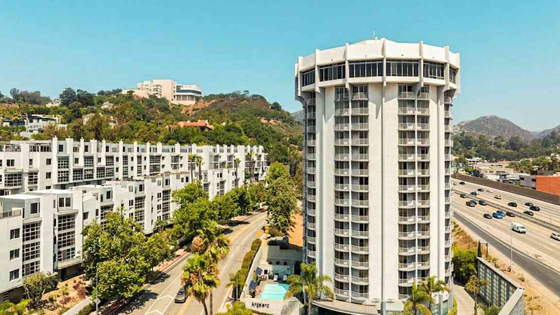 En moderne høyblokk omgitt av veier, grøntområder og andre bygninger. (AI-generert tekst)
