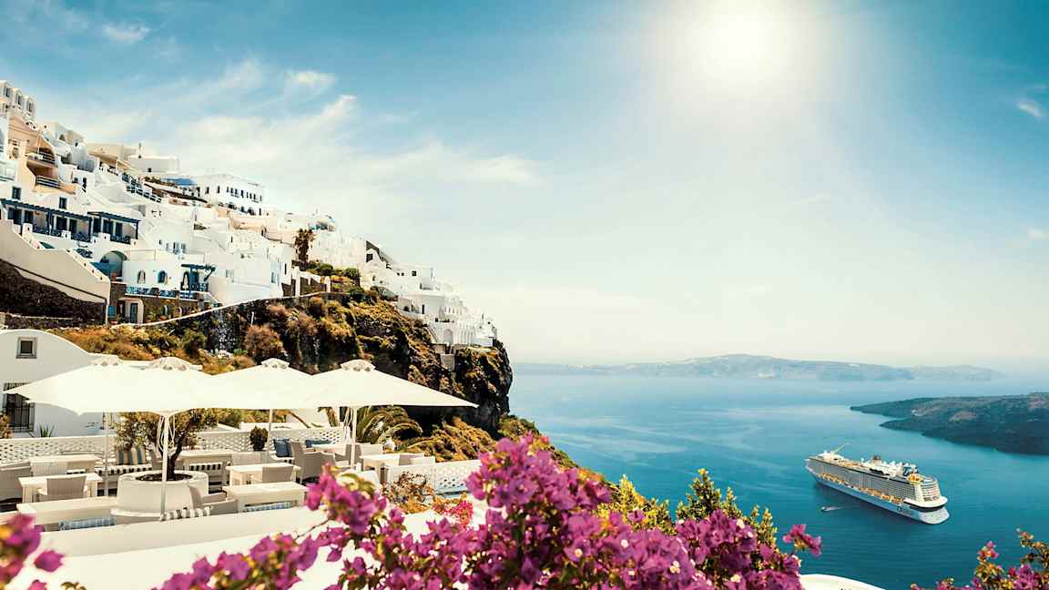 Hvite bygninger på en klippe med havutsikt og blomster i forgrunnen. Et cruiseskip vises på vannet. (AI-generert tekst)