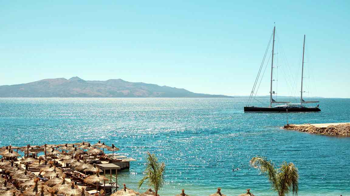 Strand med parasoller og lang seilbåt på havet med fjell i bakgrunnen. (AI-generert tekst)