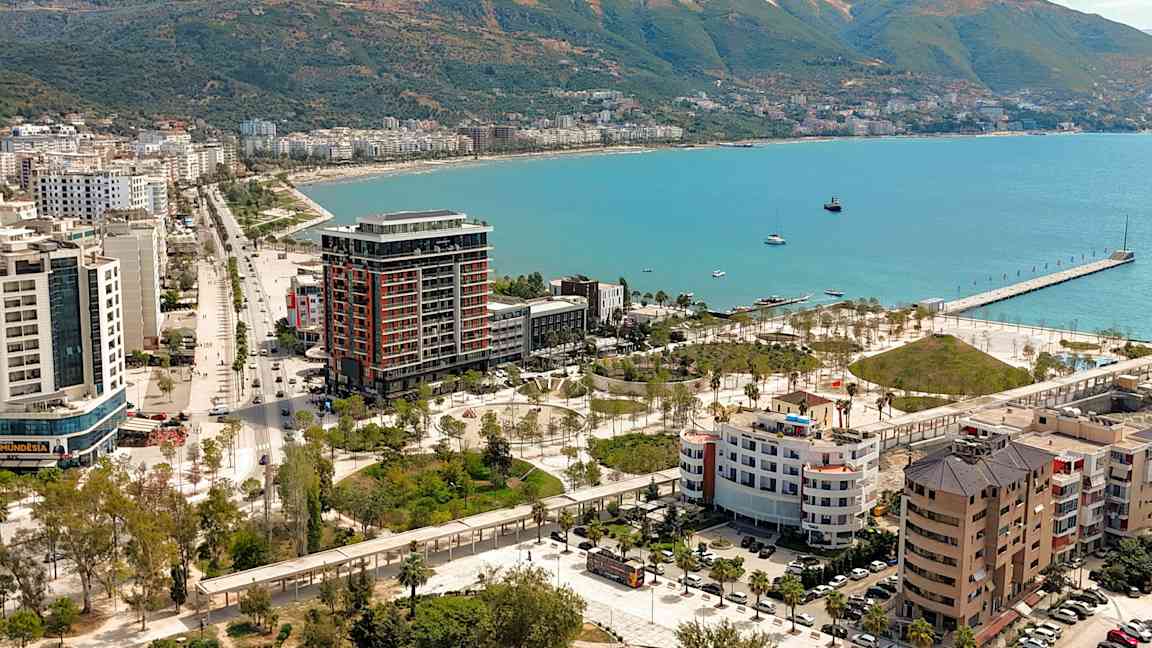 Utsikt over en kystby med bygninger, grøntområder og en brygge ved havet. (AI-generert tekst)
