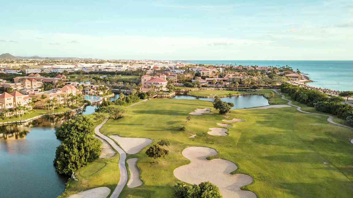 Golfbane med sandbunkere og grønne plenområder nær vannløp og strand. Bygninger i bakgrunnen. (AI-generert tekst)