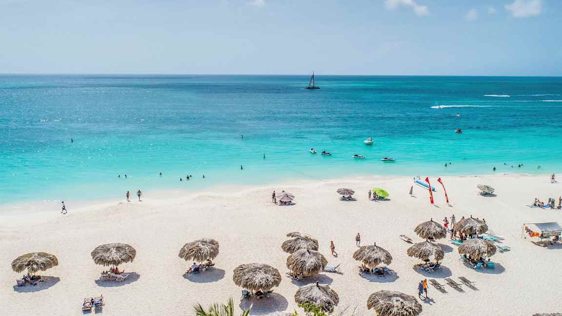 En sandstrand med parasoller og mennesker som soler seg og bader i en blå havlandskap. (AI-generert tekst)