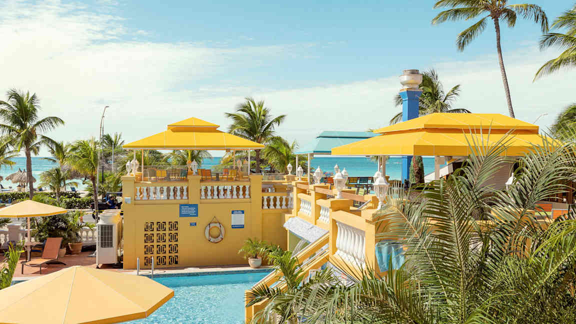 Et tropisk miljø med et basseng og gule parasoller, omgitt av palmer med havet i bakgrunnen. (AI-generert tekst)