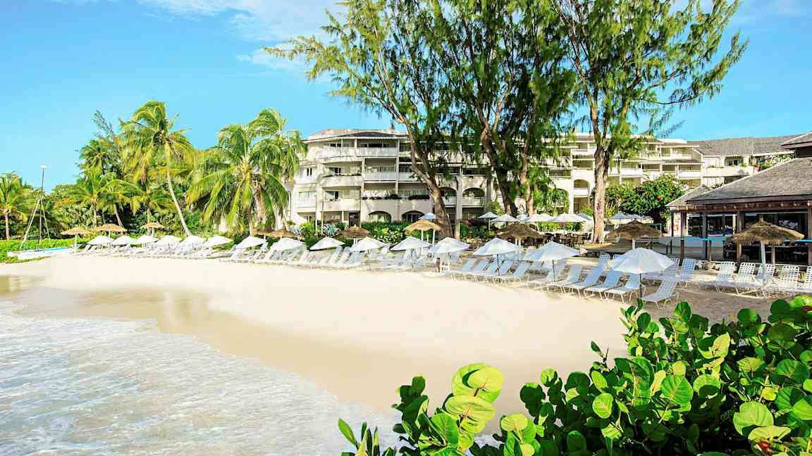 En strand med solsenger og parasoller foran en resort med palmer og beplantning. (AI-generert tekst)