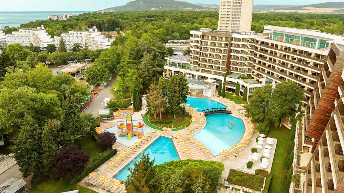 Utsikt over et hotellområde med bassenger, solsenger og omkringliggende trær. (AI-generert tekst)