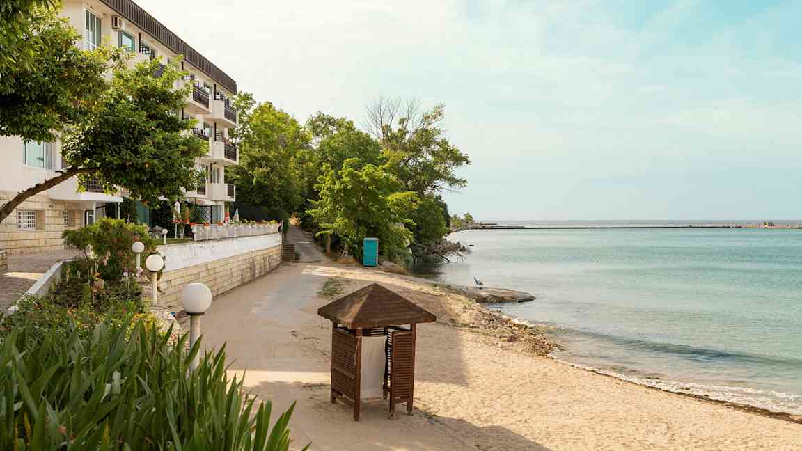 En bygning med flere balkonger ved siden av en sandstrand og hav med trær og en paviljong i forgrunnen. (AI-generert tekst)