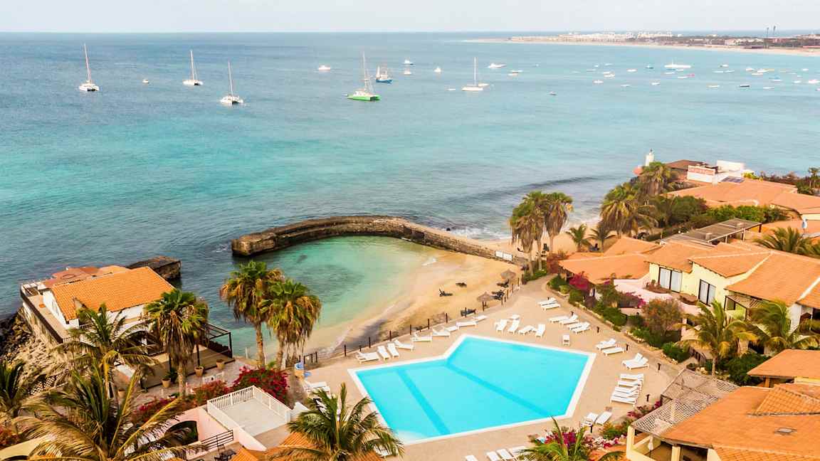 Utsikt over en badeplass og et basseng ved sjøen, omgitt av bygninger med teglrøde tak. (AI-generert tekst)