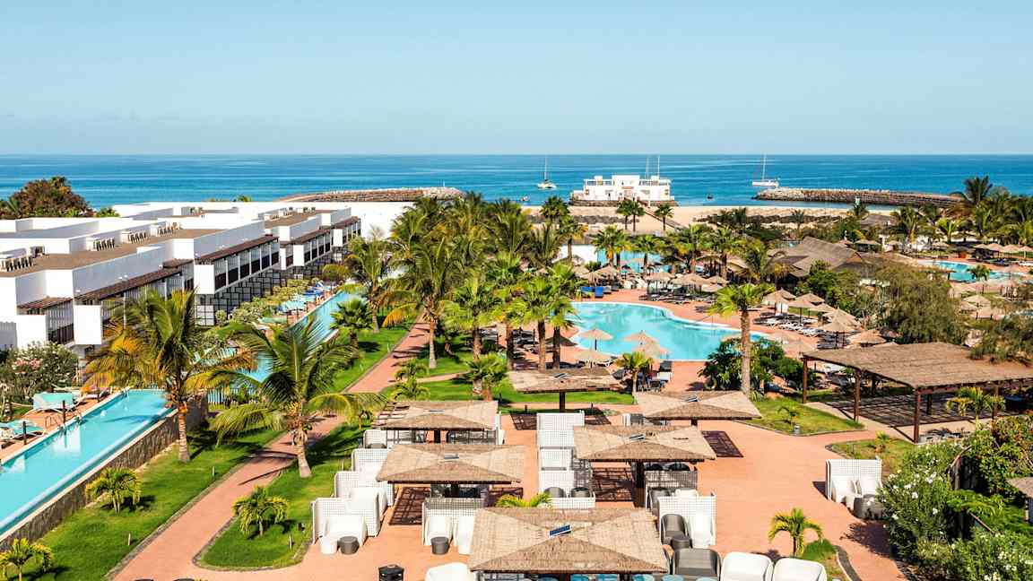 Ferieanlegg med hvite bygninger, et svømmebasseng og grøntområde rundt, samt hav i bakgrunnen. (AI-generert tekst)