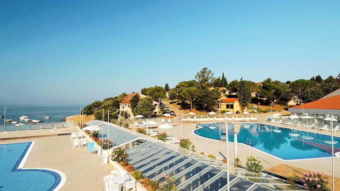 En utsikt over et utendørs svømmebasseng omgitt av en terrasse med solsenger og parasoller. I bakgrunnen er sjøen samt bygninger og vegetasjon. (AI-generert tekst)