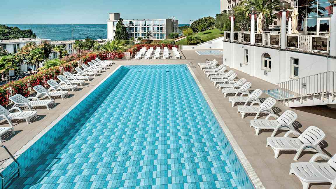 Et rektangulært basseng omgitt av solsenger, med havet og en bygning i bakgrunnen. (AI-generert tekst)