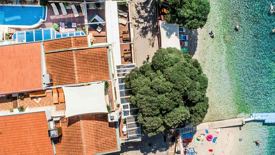 Luftfoto av en bygning med basseng og terrasse ved siden av en strand med klart vann. (AI-generert tekst)