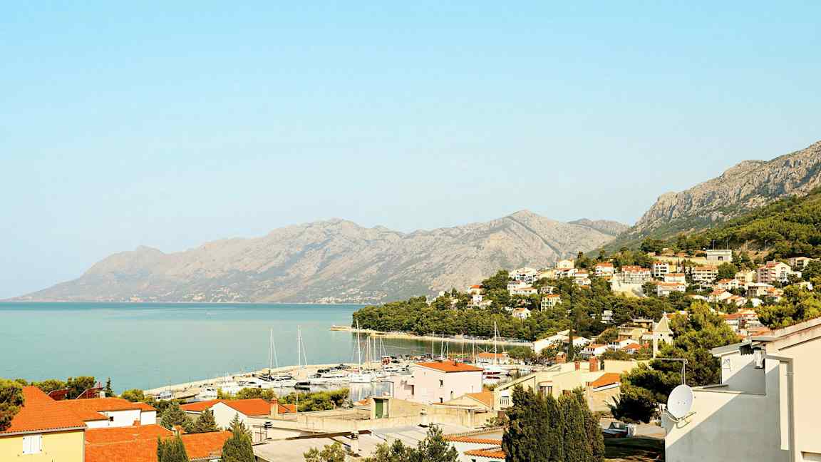 Utsikt over en kystby med røde tak, en småbåthavn og fjell i bakgrunnen. (AI-generert tekst)