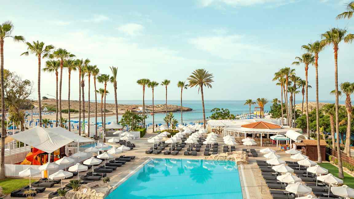 Bilde av et basseng med solsenger, parasoller og palmer ved en strand. (AI-generert tekst)