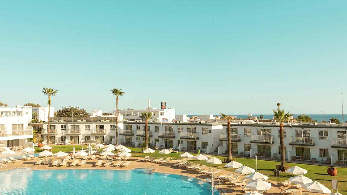 Et feriested med basseng, solsenger, parasoller og omkringliggende bygninger. (AI-generert tekst)