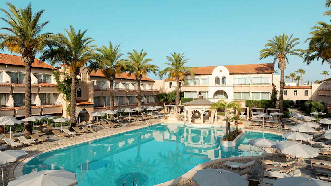 Hotellbygning med solsenger og parasoller rundt et utendørsbasseng, omgitt av palmer. (AI-generert tekst)