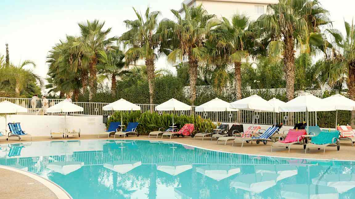 Utendørsbasseng med solsenger og parasoller omgitt av palmer. (AI-generert tekst)