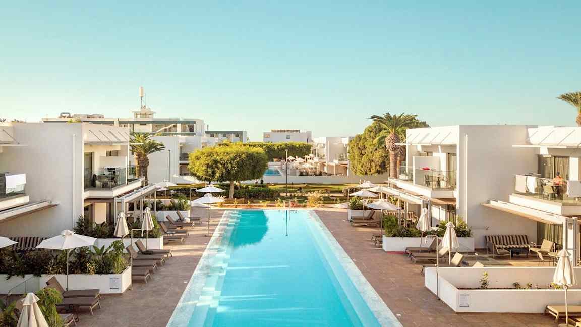 Et utendørsbasseng omgitt av moderne, hvite bygninger med balkonger og solsenger under parasoller. (AI-generert tekst)