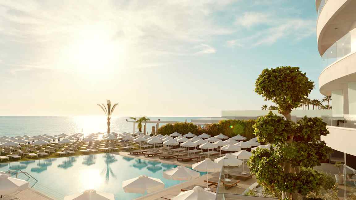 Et utendørsbasseng med solsenger og parasoller, havet i bakgrunnen. (AI-generert tekst)