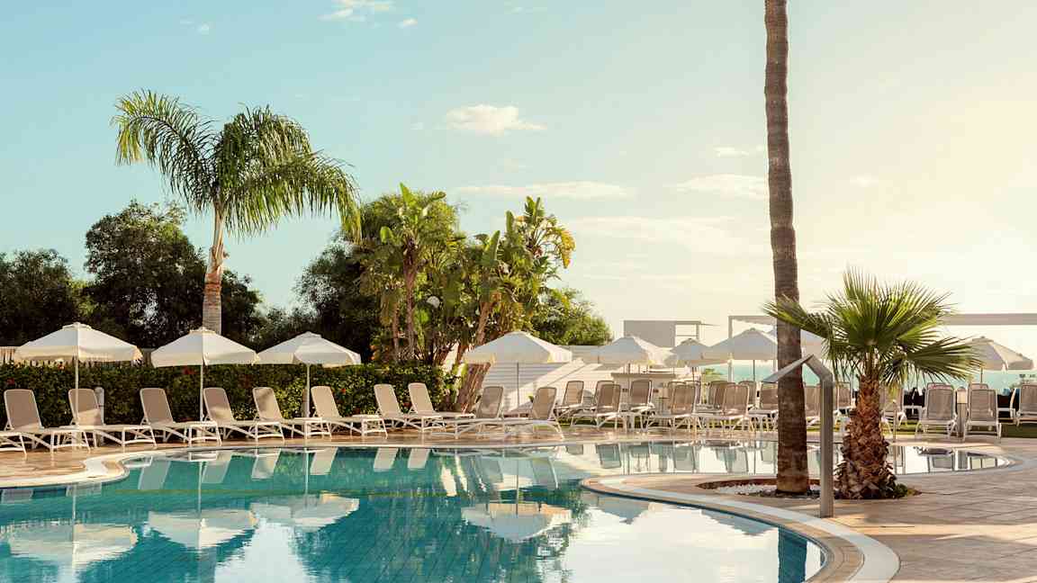 Et utendørs basseng omgitt av solsenger, parasoller og palmer. (AI-generert tekst)