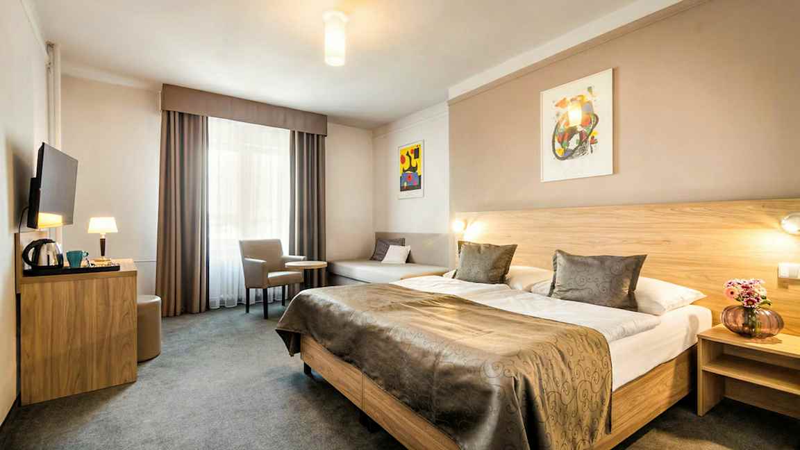 Et hotellrom med en stor dobbeltseng, et bord med to stoler og en veggmontert TV. (AI-generert tekst)