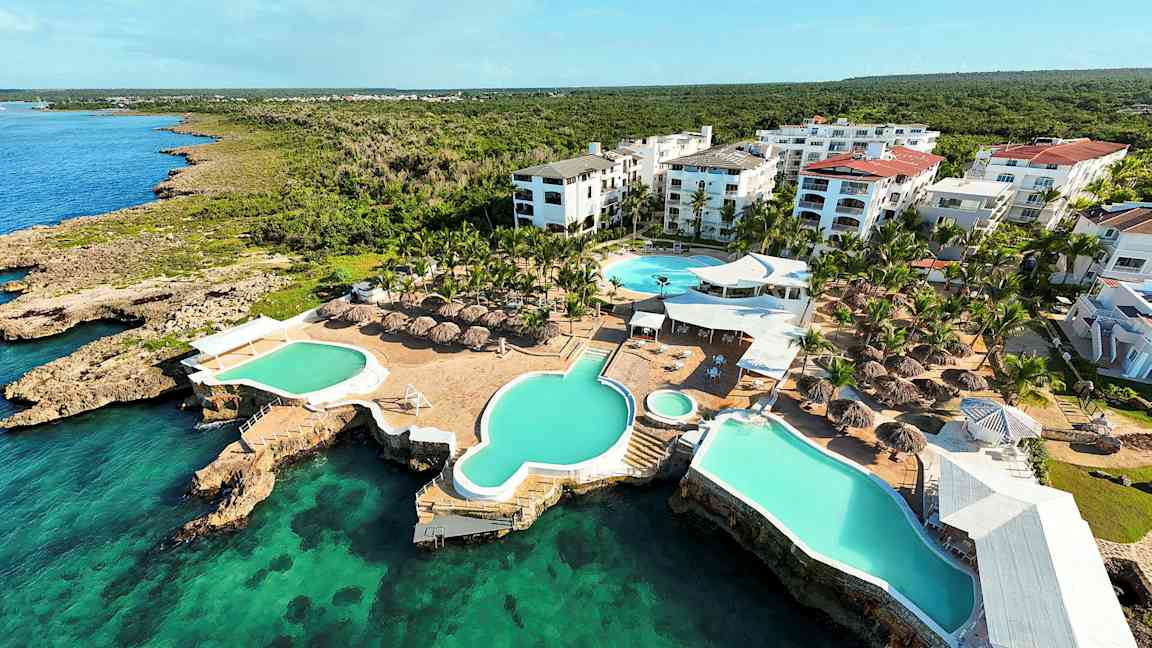 Luftfoto av et feriested med bassenger og takparasoller ved sjøen. (AI-generert tekst)