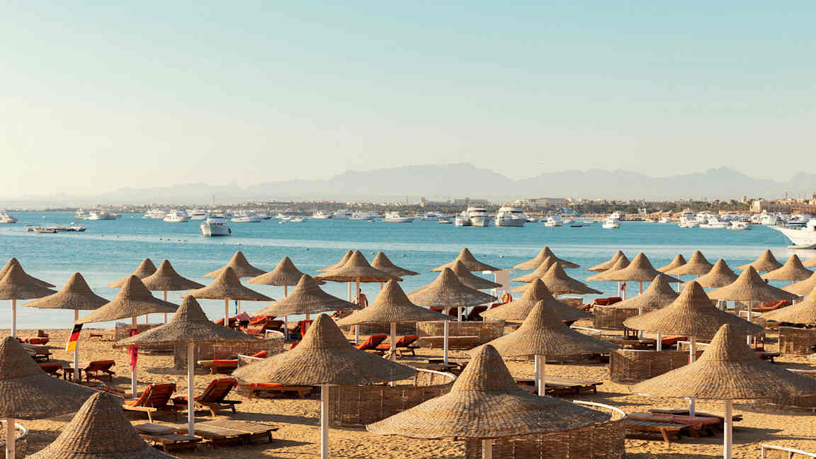 En strand med solsenger og parasoller av naturmaterialer, med sjøen og båter i bakgrunnen. (AI-generert tekst)