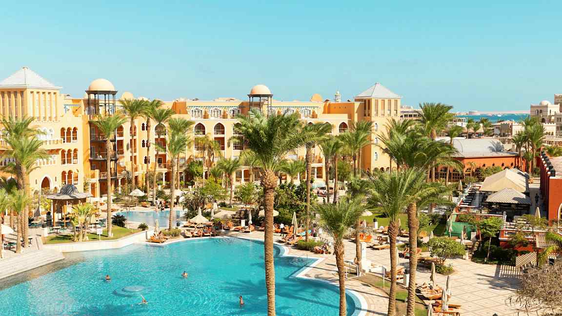 Et bilde av et hotellområde med et stort basseng, omgitt av palmer og bygninger i varme farger. (AI-generert tekst)