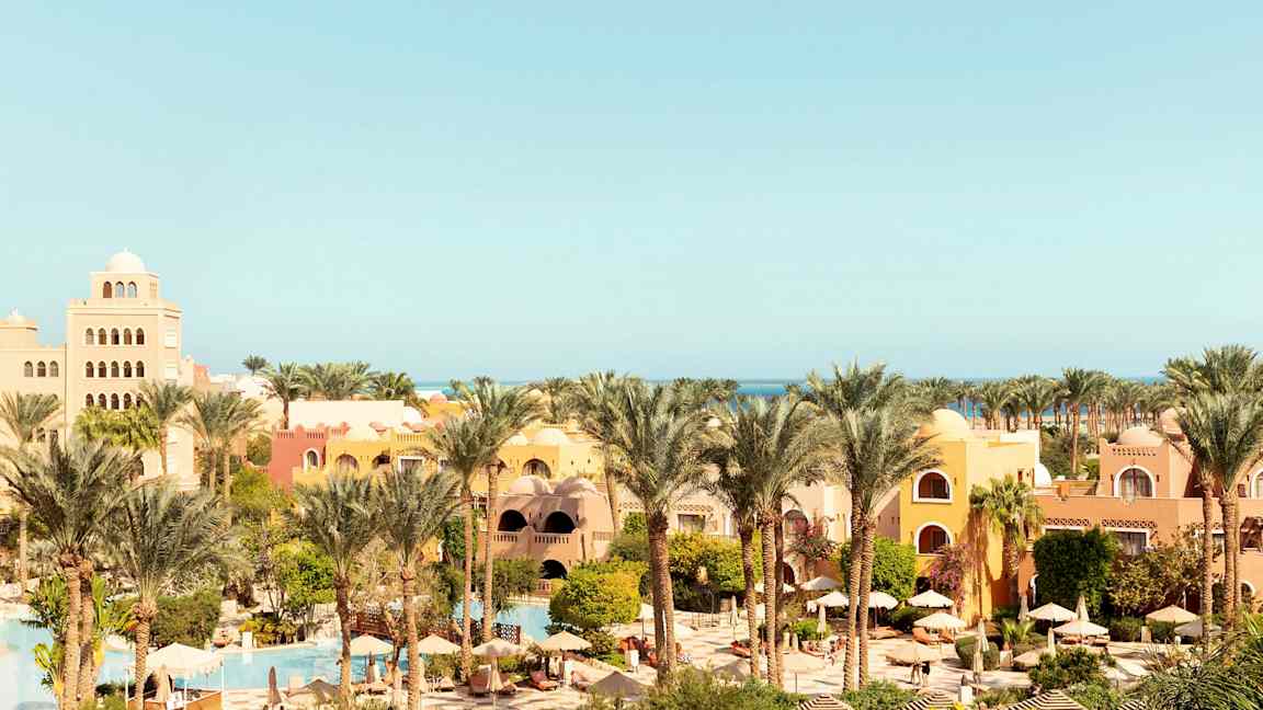 Et feriested med gule og oransje bygninger, omgitt av palmer og grønt, med bassenger og parasoller. (AI-generert tekst)