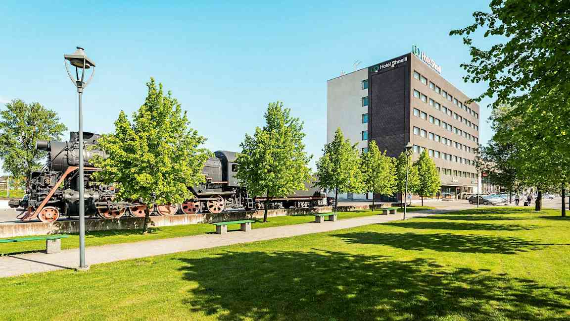 Et gammelt damplokomotiv står utendørs ved siden av et moderne hotellanlegg og frodige trær. (AI-generert tekst)